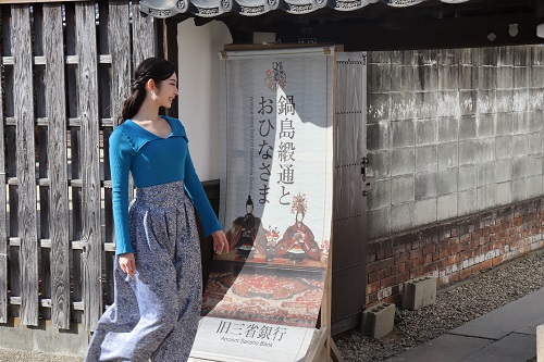 青いトップスと花柄の袴風の服を着た女性が、歴史的な街並みの建物の前にある「鍋島緞通とおひなさま」と書かれた大きな看板の横を、笑顔で歩きながら眺めている風景を捉えた写真
