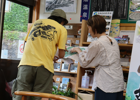 観光案内所のカウンターで、ベージュのブラウスを着た女性が黄色のTシャツを着用した男性に地図を広げて指し示しながら案内をしており、背景の棚にはパンフレットや温泉の看板が並んでいる、接客中の様子を捉えた写真