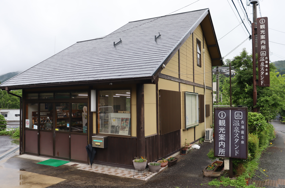 山あいの曇り空の下、ベージュの外壁と黒い屋根を持つ平屋建ての「古湯・富士観光案内所」の外観が映し出されており、建物の前には案内所と温泉スタンドを示す茶色の看板が設置されている施設写真