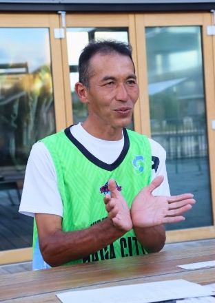 緑色のメッシュビブスを着用した藤木さんが、胸の前で両手を叩きながら、柔和な表情で話をしている様子を写した写真