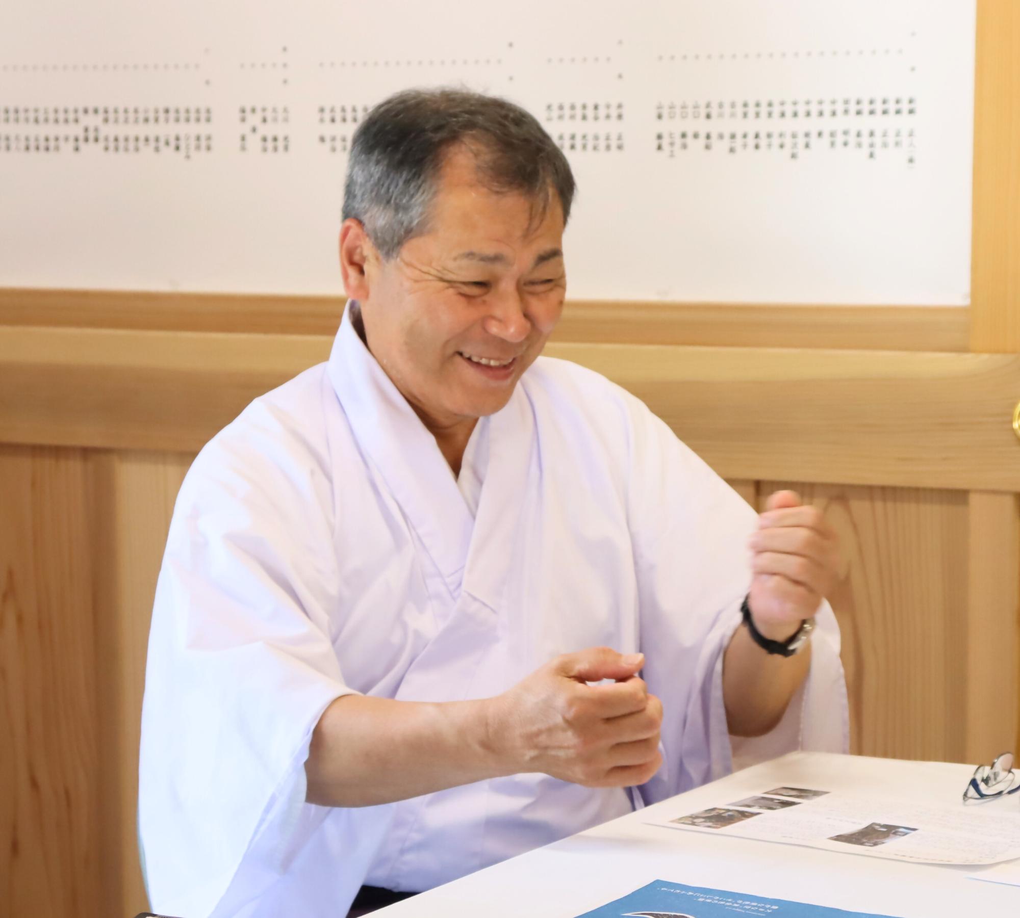 白い和装を身にまとった日吉高明さんが、明るい木目調の室内で机に向かい、身振り手振りを交えながらにこやかな笑顔で話をしている様子を捉えた写真