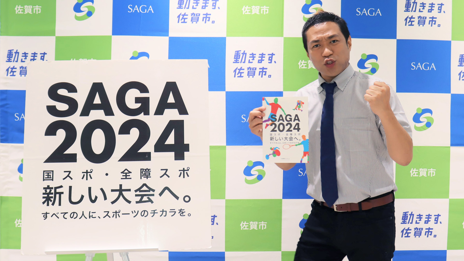 青と緑の市松模様に「SAGA」「動きます、佐賀市。」などの文字が並ぶ背景の前で、半袖シャツにネクタイ姿のはなわさんが「SAGA2024」のパネルの横に立ち、冊子を手にガッツポーズをしている写真