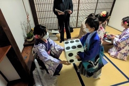 障子戸のある和室で着物姿の女性から大きなサイコロを受け取ろうとする青い袴姿の男児や周囲で見守る着物姿の女性たちを捉えた、西九州大学短期大学部の留学生と子供が交流する英会話スクールの写真