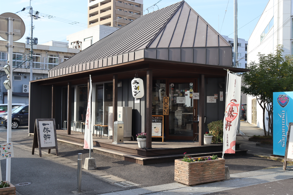落ち着いたグレーの外壁に急勾配の切妻屋根が特徴的な平屋建てのラーメン店「一休軒」の外観で、店舗前には「ラーメン」と書かれた提灯や木製のプランター、立て看板、のぼり旗が並んでいる、店舗の外観写真