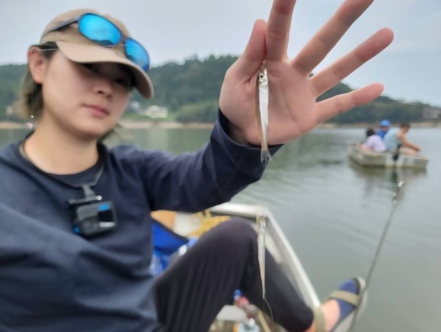 ボートに乗った女性が釣り上げたばかりの小さなワカサギを指でつまんでカメラに近づけ見せている写真