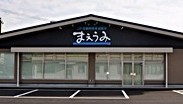 落ち着いたグレーの外壁に黒い三角形の屋根が乗り、正面中央に白抜き文字で「まえうみ」と書かれた大きな看板を掲げ、全面に並ぶ大きなガラス窓と入り口の自動ドアが特徴的な建物外観の写真（佐賀市観光協会公式ポータルサイト サガバイドットコム [sagabai.com]のサイトへリンク）