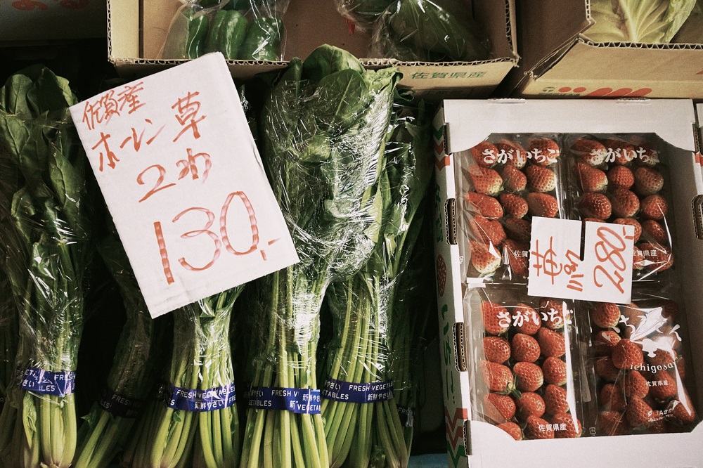 透明なラップで包まれた佐賀県産のほうれん草と、パックに入った赤く熟したイチゴが並び、それぞれに「130円」「280円」と手書きされた白い価格札が添えられている、商店で販売されている農産物の写真