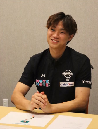 黒いポロシャツを着用した茶髪の井上 諒汰さんが、テーブルの前で両手を組み、穏やかでリラックスした表情を浮かべながら、インタビューに応じている写真