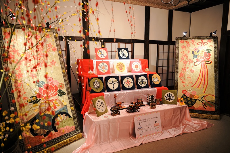 紅白の布が敷かれた雛壇に家紋や吉祥文様をあしらった色鮮やかな飾り板が並び、その左右を大きな花の刺繍パネルと餅花が彩る、モダンで芸術的な雰囲気を持つ雛飾りの展示風景の写真