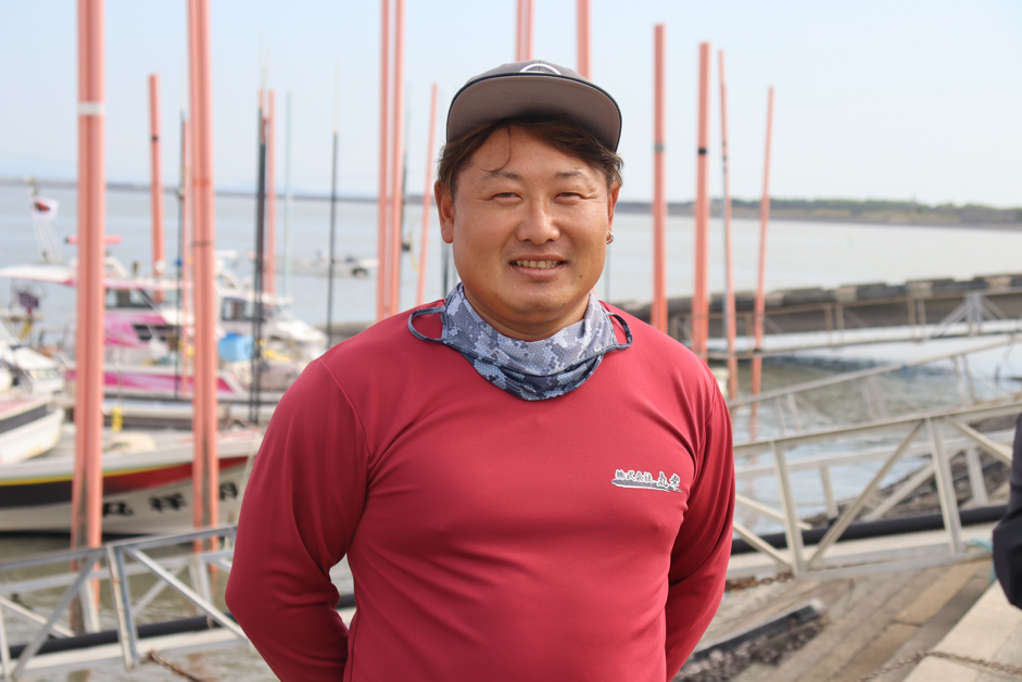 漁港の岸壁を背景に、グレーの帽子と赤い長袖シャツを身につけた竹下幸佑さんが笑顔で写っている写真