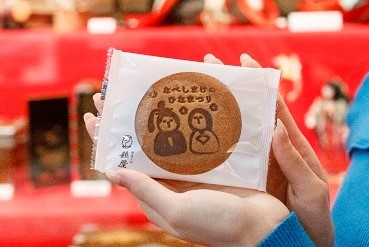 赤い雛壇を背景に、かわいらしい内裏雛のイラストとともに「なべしまけのひなまつり」という文字が刻印されている袋入りの丸い煎餅を両手で持った女性の手元をクローズアップで捉えた写真