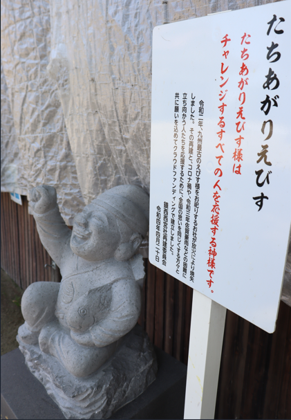 右拳を力強く突き上げて満面の笑みを浮かべる「たちあがりえびす」の石像の横に、その由来と応援のメッセージが記された白い案内看板が立てられており、背景には銀色のシートが張られている写真