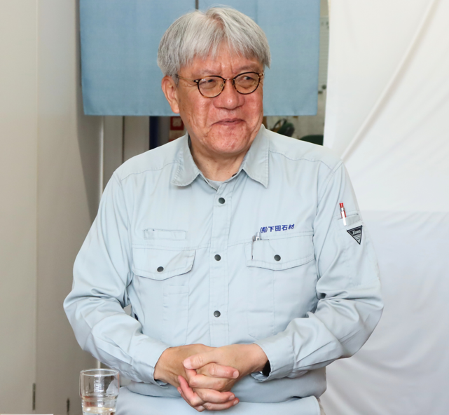 背景には水色の暖簾や白い布がシンプルに配置されている前で、薄いブルーの作業服を着用し眼鏡をかけた白髪交じりの男性が、胸元で両手を組みながら柔らかな微笑みを浮かべて立っている写真