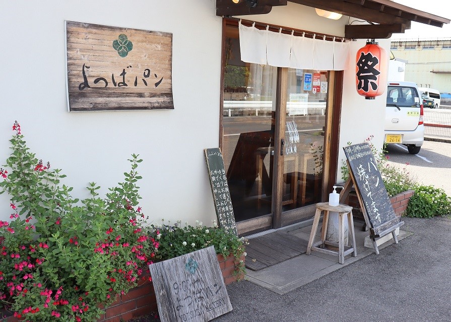 白壁に「よつばパン」と記された木製看板が掲げられ、入り口には白い暖簾と「祭」と書かれた赤い提灯が吊るされ、足元には色鮮やかなピンクの花が咲くプランターや木製の案内板が置かれた店舗外観の写真。