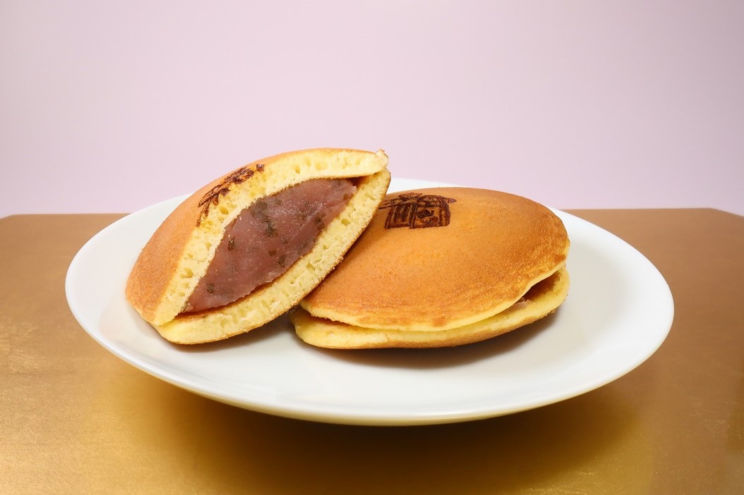 ふっくらと焼き上げられた黄金色の生地に可愛らしい焼印が押され、中にはしっとりとした粒あんがたっぷりと詰まったどら焼きが、白い平皿の上で断面を見せるように盛り付けられた和菓子の写真