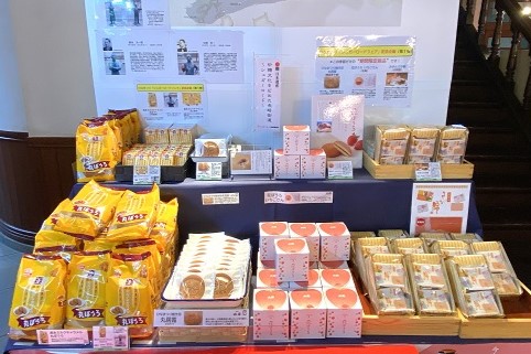 歴史を感じる木造建築の店内で、黄色いパッケージの丸ぼうろや、ピンク色の個包装が積み上げられた、佐賀県を代表する銘菓が「シュガーロードフェア」の看板と共に整然と並ぶ、多彩な特産品が揃ったお土産売り場の写真