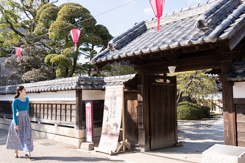 瓦屋根の立派な門と白壁が続く日本情緒豊かな建物の前を、青いトップスと花柄のロングスカートを身にまとった女性が晴天の下で優雅に歩いており、門の脇にはイベントの立て看板が置かれている穏やかな風景写真