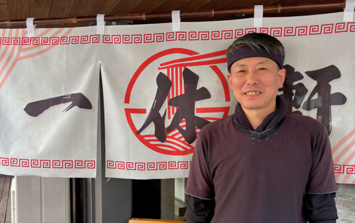 「一休軒」と書かれた暖簾の前で、黒いシャツにバンダナを巻いた店主・時吉学さんが優しく微笑んでいる写真