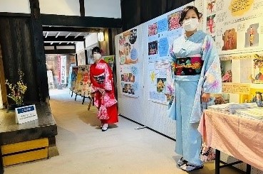 歴史ある和室の展示会場で、水色の着物を着た女性と赤い着物の女性が、海外イベントや日本文化の功績を紹介する数多くの解説パネルが並ぶ通路に立ち、来場者を案内するように佇んでいる様子の写真