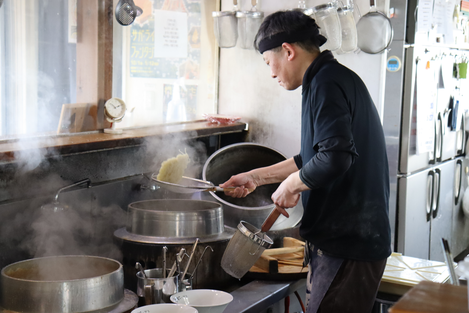 湯気が立ち込める厨房で、店主の時吉さんが平ザルを使って大きな釜から茹で上がった麺を鮮やかに湯切りしている調理風景を捉えた写真