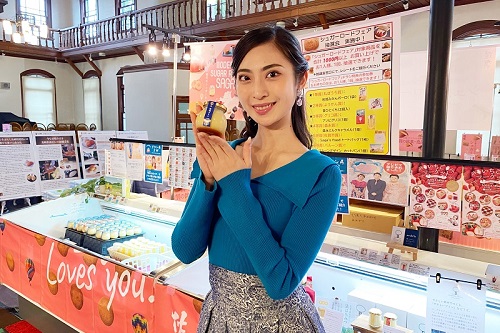 青いトップスを着た松尾綺子さんが瓶入りのプリンを手に微笑んでおり、背後には「Saga Loves you!」と書かれた赤い布が掛かる冷蔵ケースに、多くのプリンが並んでいるシュガーロードフェアの様子の写真