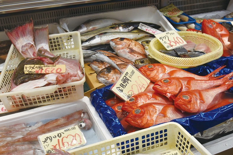 鮮やかな赤色の金目鯛や銀色のサワラ、ヤリイカなどの新鮮な魚介類が、手書きの値札とともにプラスチックの籠や発泡スチロールに整然と陳列されている、市場の活気が伝わる鮮魚店の売り場の写真