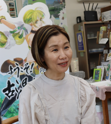 明るいベージュのブラウスを着用した茶髪のショートヘアの村岡夏子さんが穏やかな表情で微笑んでおり、背景にはアニメ風のキャラクターパネルや案内標識、棚が並ぶ室内が映り込んでいる写真
