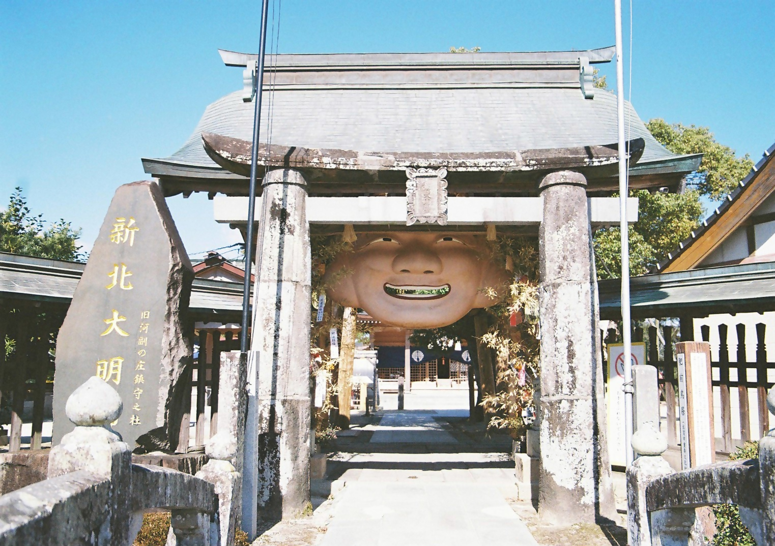 新北神社の石造りの鳥居の中央に、にこやかな表情を浮かべた巨大なお多福の面が飾られており、左側には「新北大明神」と刻まれた大きな石碑が立っている、伝統とユーモアが融合した外観写真