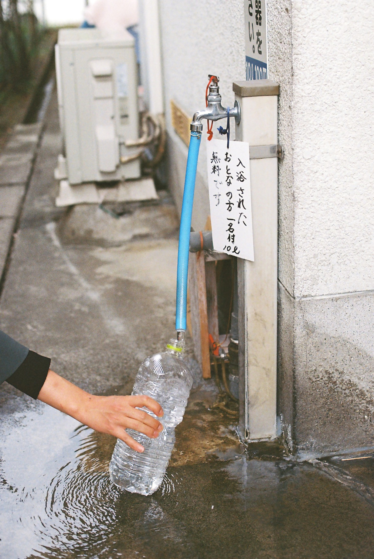 屋外の蛇口から伸びる青いホースから、透明な大きなペットボトルに水が注がれており、その傍らには「入浴されたおとなの方一名付10ℓリットル料です」と書かれた手書きの案内札が吊り下げられている、温泉スタンドでの給水の様子を捉えた写真