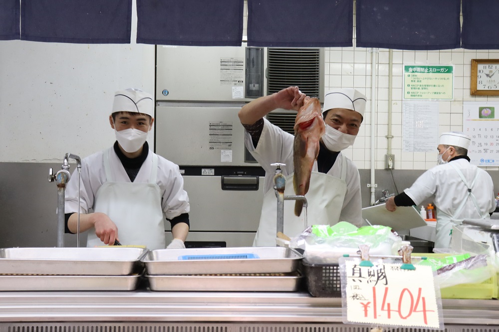 鮮魚店の調理場で、白い衛生服とマスクを着用した店員が、銀色のトレイや蛇口が並ぶカウンター越しに、大きな魚を高く掲げて見せながらカメラに向かって微笑んでいる、活気ある作業風景の写真