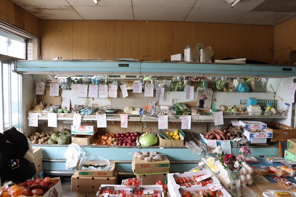 商品の並ぶ商店の棚に、ジャガイモ、カボチャ、玉ねぎ、キャベツ、レモンなどの多様な野菜や果物が段ボール箱ごとに整然と並べられ、上部の棚には商品の名称と価格が記された多くの白い紙が貼られている店内の写真