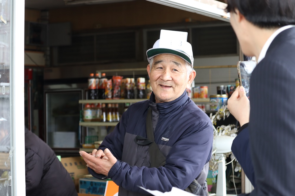 商品の並ぶ商店の軒先で、紺色のジャンパーと白い帽子を着用した中島商店代表の池田直さんが、スマートフォンを向けるスーツ姿の人物に対し、穏やかな表情で手のひらを合わせながら話をしている光景の写真
