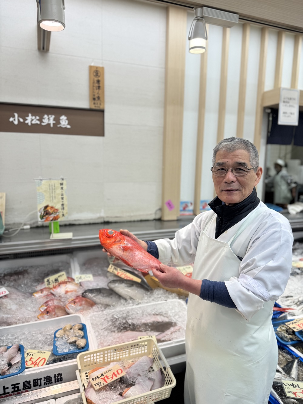 明るい店内で白い衛生服とエプロンを着用した小松鮮魚店店主の小松忠雄さんが、氷が敷き詰められた鮮魚の陳列棚を背に、立派な赤い金目鯛を両手で水平に掲げながら穏やかな表情で正面を見据える姿を捉えた写真