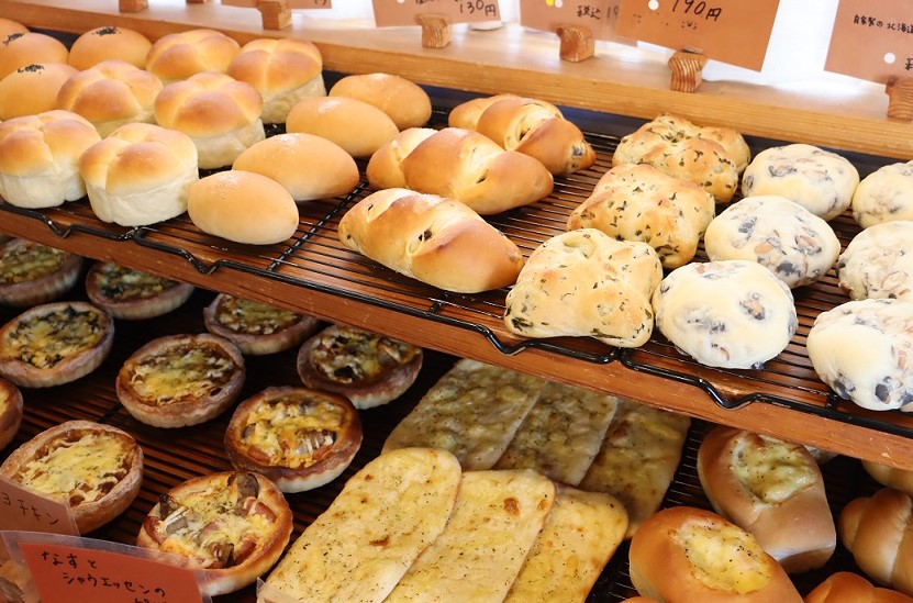 店内の木製棚に、惣菜パンや菓子パン、豆がふんだんに練り込まれた白いパンなど、多種多様な焼き立てパンが黒いワイヤーバスケットの上に所狭しと並べられ、上部には手書きのプライスカードが添えられた陳列棚の写真。