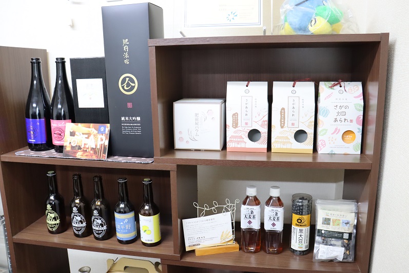 木製の棚に、地元の麦を使用した二条大麦茶のペットボトルやクラフトビール、日本酒の瓶、そして可愛らしいイラストが描かれた「さがあられ」などの加工食品が整然と並べられている写真