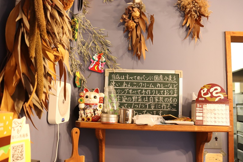 店内の薄紫色の壁面を背景に、ドライフラワーや招き猫が飾られた木製棚の上で、すべてのパンに国産小麦を使用している旨が丁寧に手書きされた小さな黒板と、2025年のカレンダーが並ぶ店内の装飾写真。