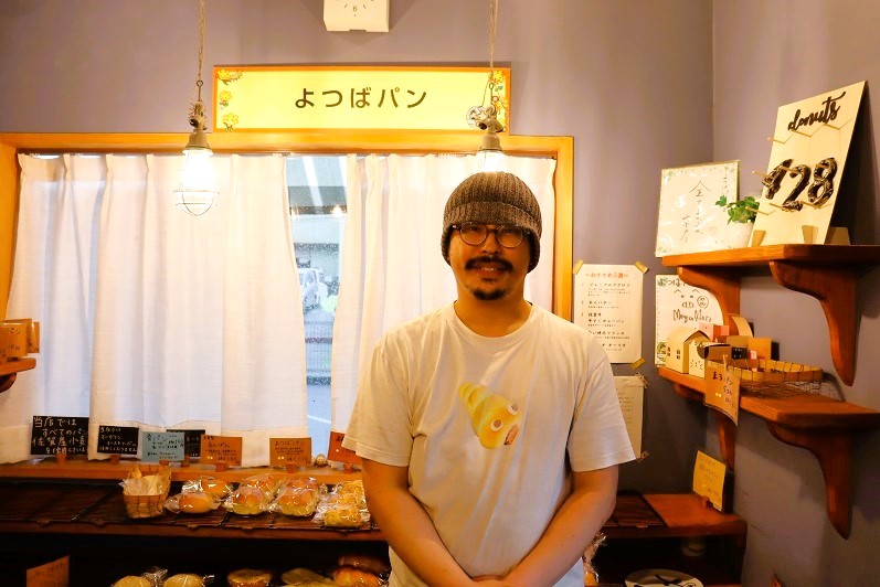 「よつばパン」の店内で、グレーのニット帽と眼鏡を着用した淵慶介さんが、コロネが描かれた白いTシャツを着て、温かな照明の下で穏やかな笑みを浮かべて正面を向いて立つポートレート写真。
