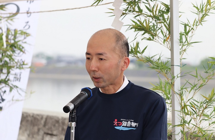 「えつ銀色祭り」のロゴが入った紺色のシャツを着用した吉田宏毅会長が、屋外の川沿いに設置されたマイクの前で、竹の装飾を背に真剣な表情で挨拶を行っている写真