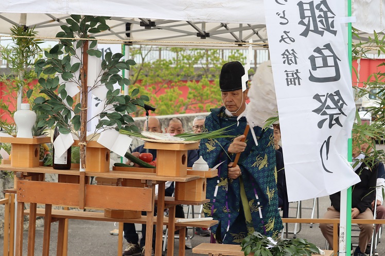 屋外のテント下で、青い装束を纏った神職が白い大幣を振り、野菜や魚が供えられた神棚の前で儀式を執り行う、佐賀市もろどみin食の会の会員らが見守る中で行われた「えつ」の豊漁祈願祭の様子を捉えた写真