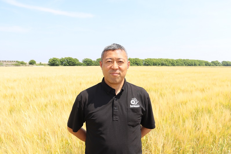 強い日差しが降り注ぐ広大な黄金色の麦畑を背景に、左胸に白いロゴがプリントされた黒いポロシャツを着用した池田大志さんが、穏やかな表情で正面を見つめて立っているポートレート写真。