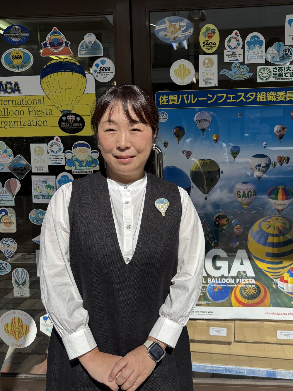 熱気球のブローチを胸に付けたにしくぼさんが、歴代の大会ステッカーやポスターが貼られたガラスドアの前に立ち、穏やかな微笑みを浮かべている写真
