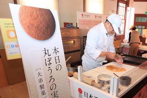 白いキャップと調理服を身につけた職人が、平らな作業台の上で丸ぼうろの生地を丁寧に整えており、手前には「本日の丸ぼうろの実演」と書かれた大きな看板が立てられた、シュガーロードフェアでの調理実演風景の写真