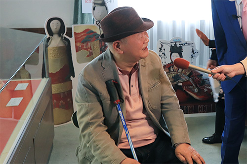 茶色のハットとベージュのジャケットを着用し杖を手にした大塚清吾さんが、展示ケースや雛人形のパネルが置かれた会場でマイクを向けられ、江戸時代から伝わる貴重な「次郎左衛門雛」について穏やかな表情で解説している写真