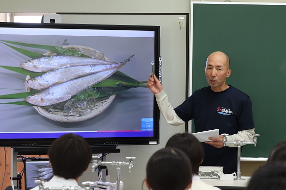 教室内で男性が大型モニターに映し出されたザルに乗った3匹の魚の画像を指差しながら、手元の資料を確認しつつ生徒たちに向かって説明を行っている授業風景を捉えた写真