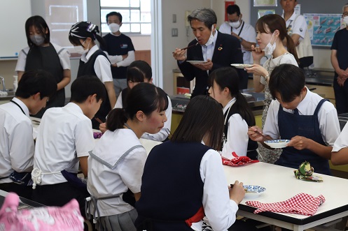 明るい調理実習室にて、エプロンを身に着けた生徒たちが机を囲んで座り、自分たちで調理した魚料理を箸を使って静かに試食している一方で、後方では教員や関係者と思われる大人たちも一緒に味を確かめている、和やかな試食風景を捉えた写真