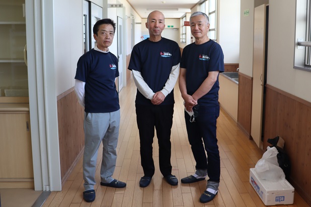 学校の廊下にて、お揃いの紺色のロゴ入りTシャツを着用した3人の男性が横一列に並び、カメラに向かって穏やかな表情で立っている写真