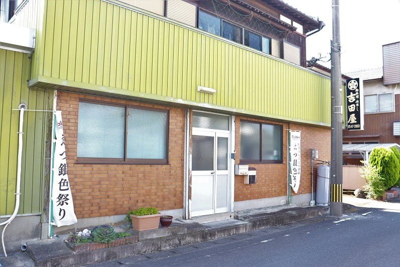 明るい黄色の外壁と茶色のレンガ壁が特徴的な二階建ての建物の外観で、入口の左右には「えつ銀色祭り」と書かれた白い幟が立てられ、右側の電柱には「吉田屋」と記された看板が設置されている店舗正面の写真