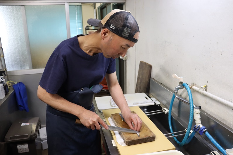 黒とベージュのメッシュキャップに紺色のTシャツ、エプロンを身に着けた吉田 宏毅さんが、厨房の流し台の前で木製のまな板の上に置かれた細長い魚に真剣な表情で包丁を入れ、丁寧にさばいている様子を横から捉えた写真