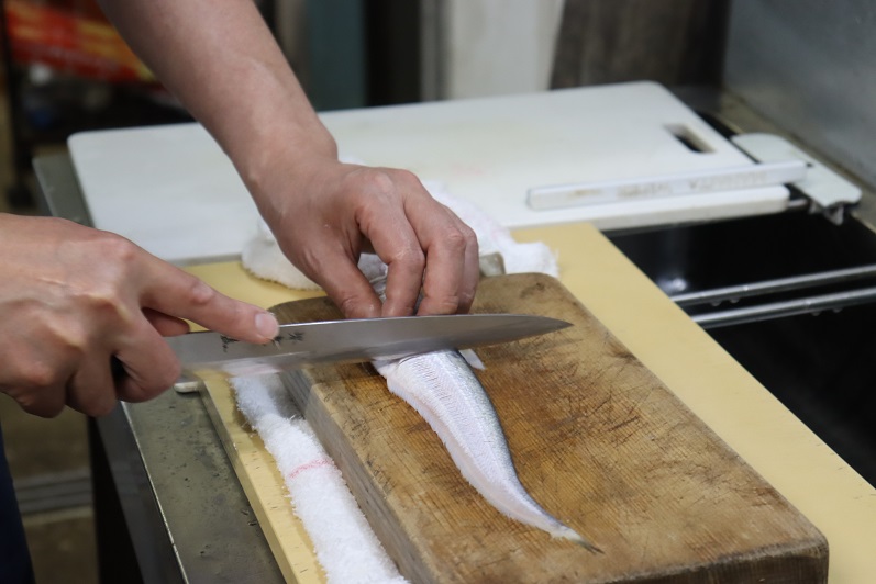 木製のまな板の上に置かれた細長く銀色の魚に対し、料理人が左手で魚体を固定しながら右手に持った鋭い刺身包丁で、背に沿って慎重かつ鮮やかに包丁を入れ、三枚おろしの工程を進めている手元の様子を克明に捉えた写真