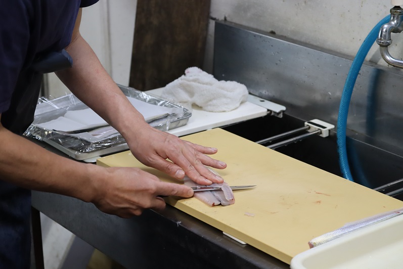 まな板の上に広げられた魚の身を左手で優しく押さえながら、料理人が右手の包丁を水平に滑らせて薄く切り分ける繊細な作業を行っており、背後には清潔に保たれた厨房のシンクが見える、職人の熟練した手技を捉えた写真
